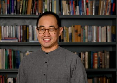 Person in gray shirt with glasses standing in front of bookshelf.