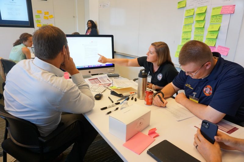 Three men and women sit at table during a discussion about artificial intelligence. One points to a computer screen with information displayed on it.