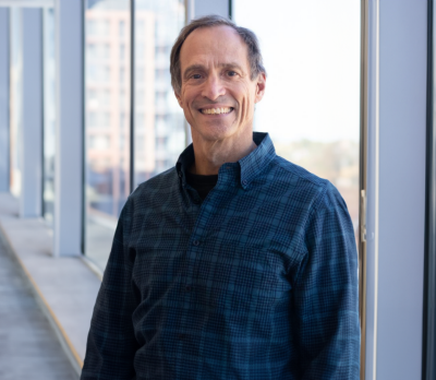 A man, wearing a blue plaid shirt, stands in front of a window, smiling. 