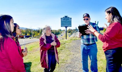 Katie Lee, Sadhana Manthapuri, and Elli Travis meet with visitors, standing in front of a Battle of New Market sign and rustic fences.