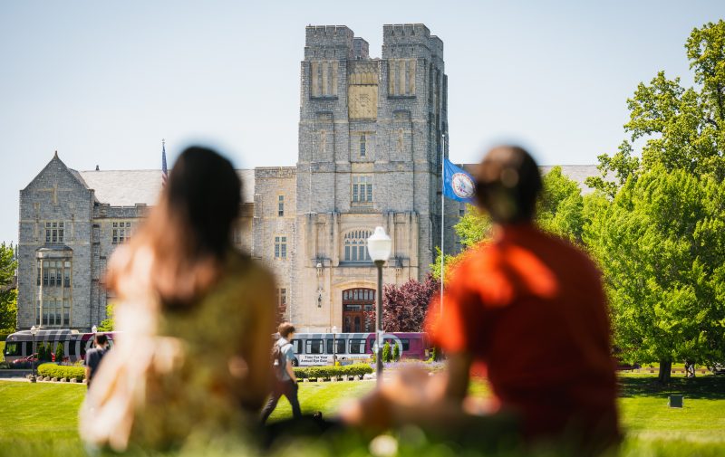 Two students in the foreground sitting beneath a tree on the Drillfield near Burruss Hall